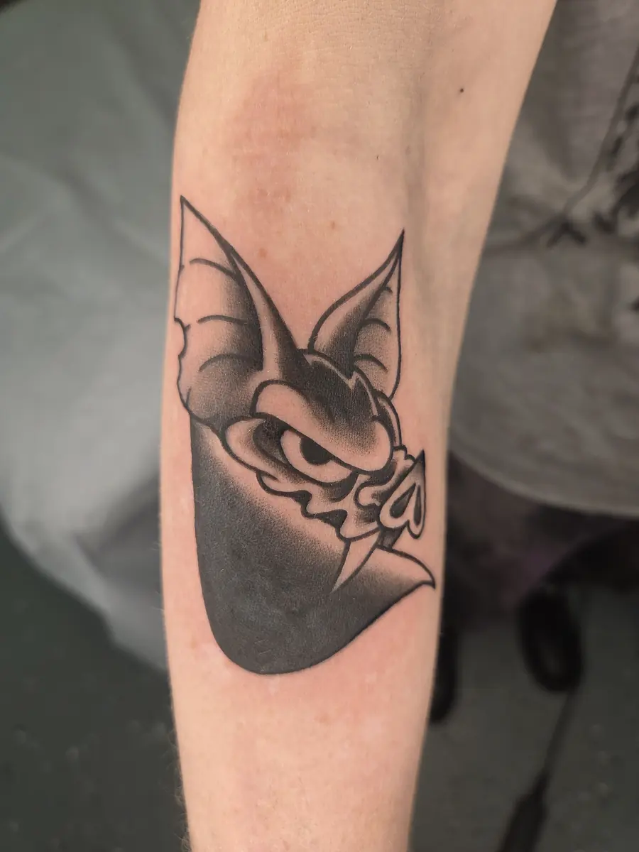 Black and grey cartoon bat character tattoo on a forearm