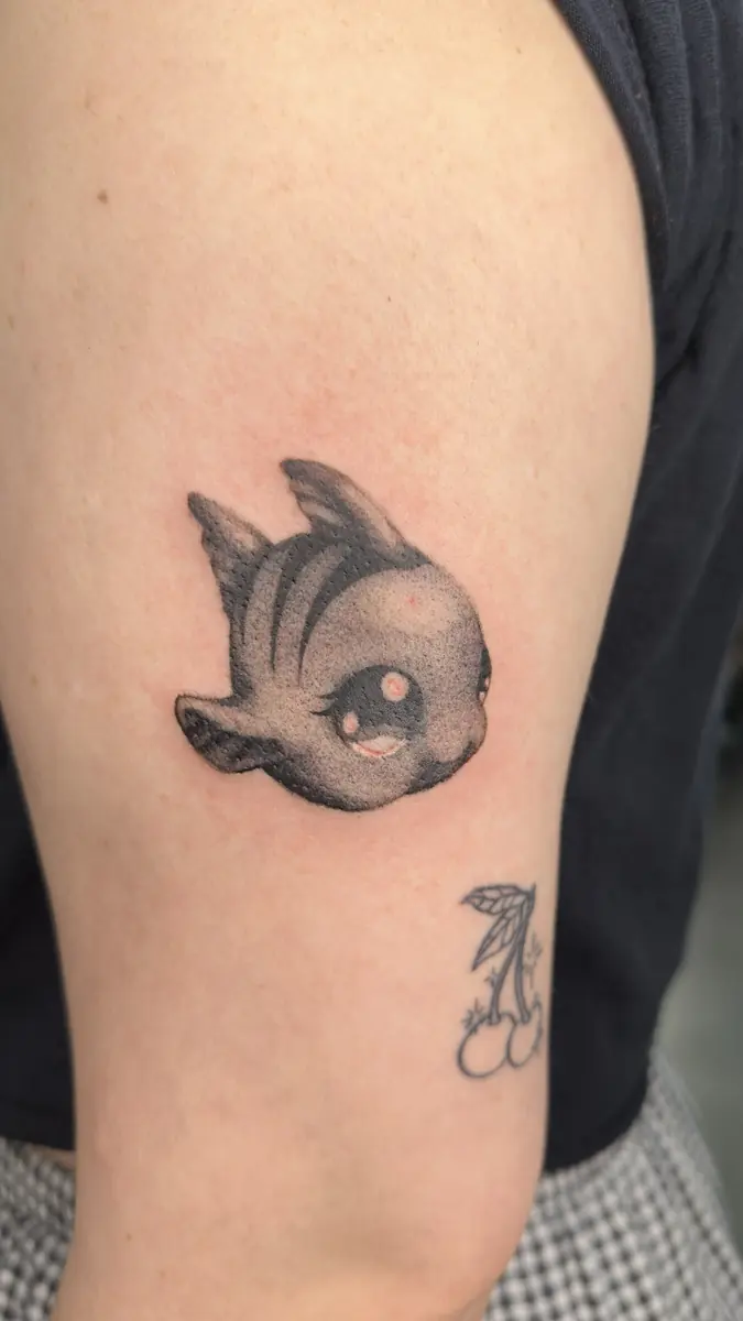 Black and grey cartoon fish character tattoo on an upper arm