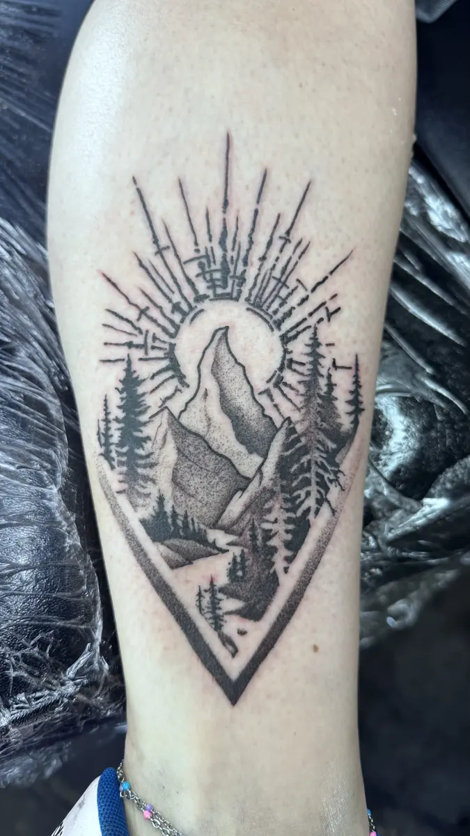 Blackwork mountain and pine landscape tattoo inside a geometric frame with sun rays, on a calf
