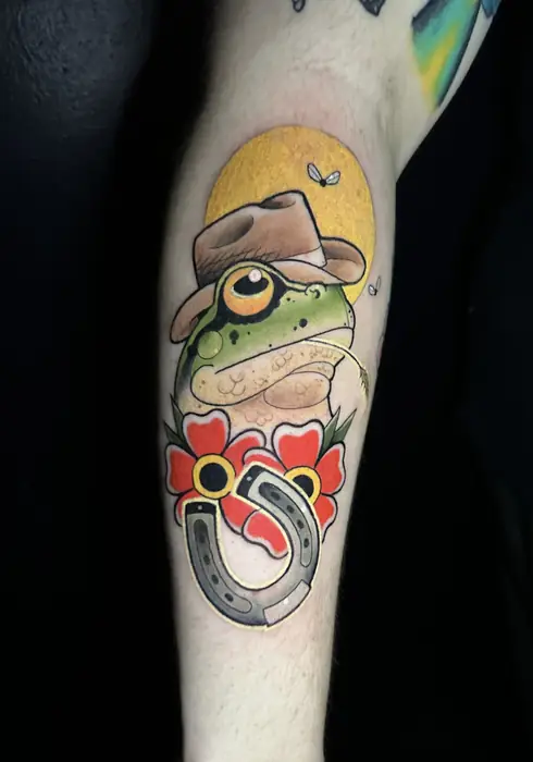 Neo-traditional tattoo of a frog in a hat resting on a lucky horseshoe with red flowers, tattooed at Blind Fox Tattoo in Amarillo, TX