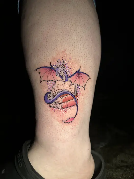 Watercolor style tattoo of a purple dragon rising from an open book on an ankle