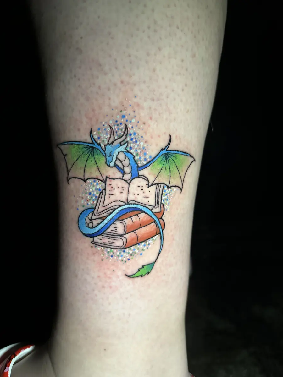 Watercolor style tattoo of a green dragon rising from an open book on an ankle